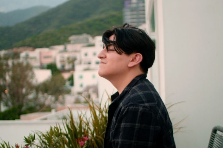 Person with dark hair and plaid shirt faces a mountain view from a balcony.