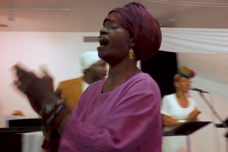 A woman in a purple top and head wrap sings and claps. Other singers are behind her.