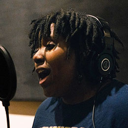 Woman with dreadlocks singing into a studio microphone.