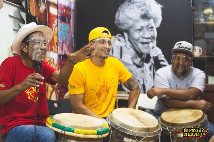 Three musicians with face shields play drums and speak into a microphone.  A large black and white photograph of a singer hangs on the wall behind them.