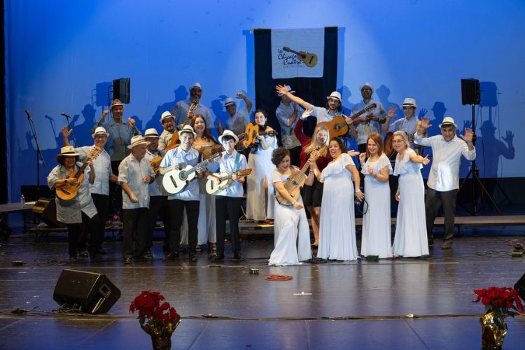 A group of musicians onstage smile and wave, holding various stringed instruments. A banner behind them reads "Chicago Cuatro."