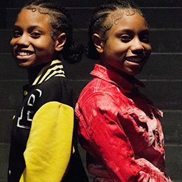 Smiling twins pose back-to-back, one wearing a letter jacket.