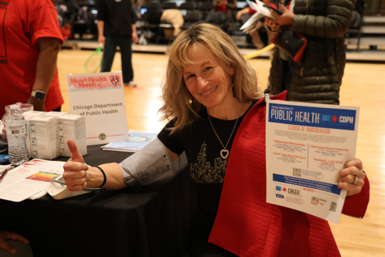 Smiling woman with blood pressure cuff, giving a thumbs up, holding a public health flyer.