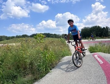 A smiling boy in a helmet rides a red BMX bike on a paved pump track surrounded by nature on a sunny day.
