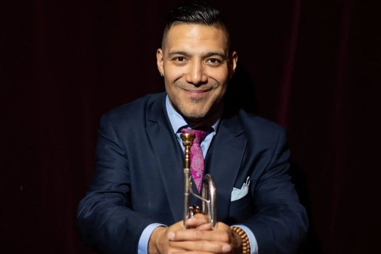 Photograph of a smiling man in a suit and purple tie, holding a trumpet against a dark background.