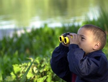 A young boy in a blue jacket looks through bright yellow toy binoculars outdoors.