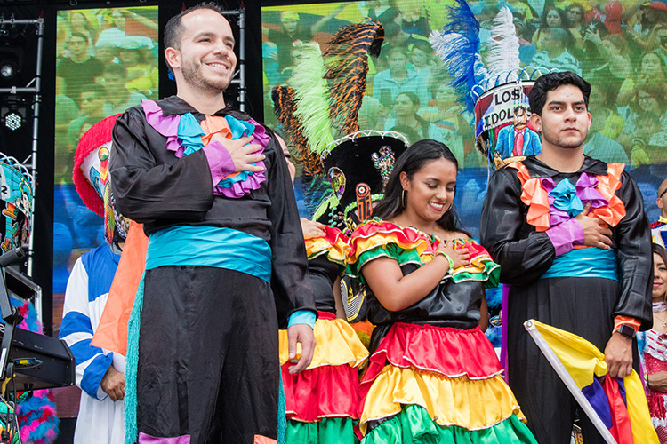 People in colorful folk costumes standing on a stage with hands over their hearts.