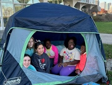 Group of children smiling inside a dark blue and grey Coleman tent outdoors.