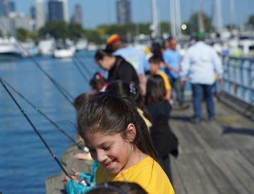 Photograph of a group of children fishing on a wooden pier with a city skyline in the background.