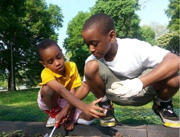 Two boys crouching in a park, working together to garden in a dirt plot.