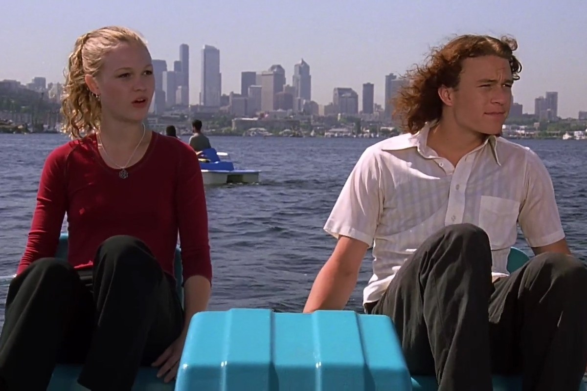 Julia Stiles and Heath Ledger in a paddle boat on the water with a city skyline in the background.