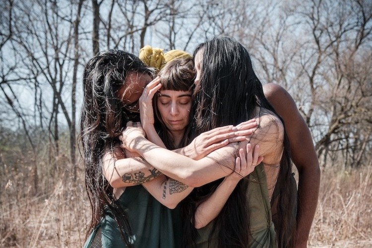 Three women with long dark hair embrace in a field of dry brush against a backdrop of bare trees.