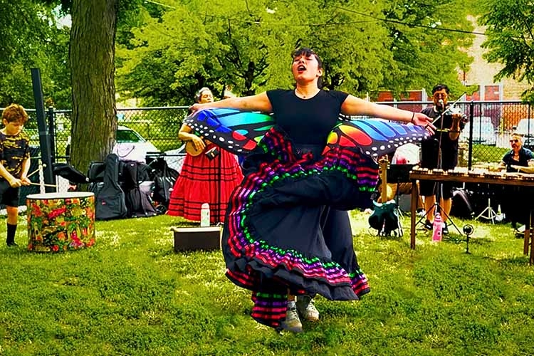 Dancer in a colorful skirt performs on a grassy lawn with musicians in the background.