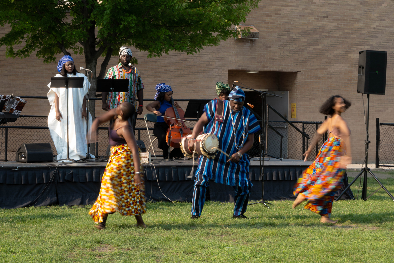 Photograph of a musical ensemble and dancers in traditional West African attire performing outdoors.