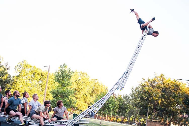 A person performs a handstand on top of a tall, precarious ladder balanced by others.