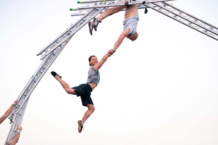 Photograph of two aerial performers suspended from a metal frame holding hands.