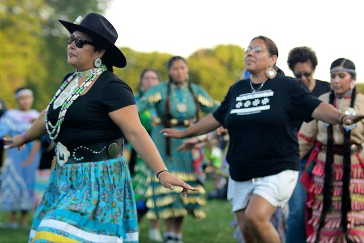 Indigenous women dancing outdoors in traditional and casual clothing.