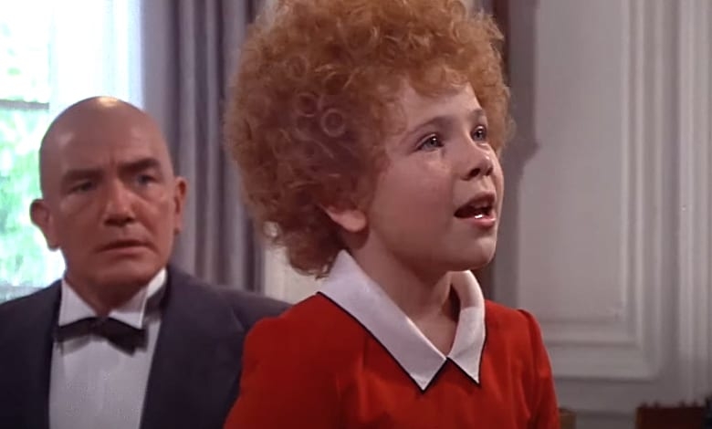 A young girl with curly red hair looks upward as a bald man in a tuxedo watches her.
