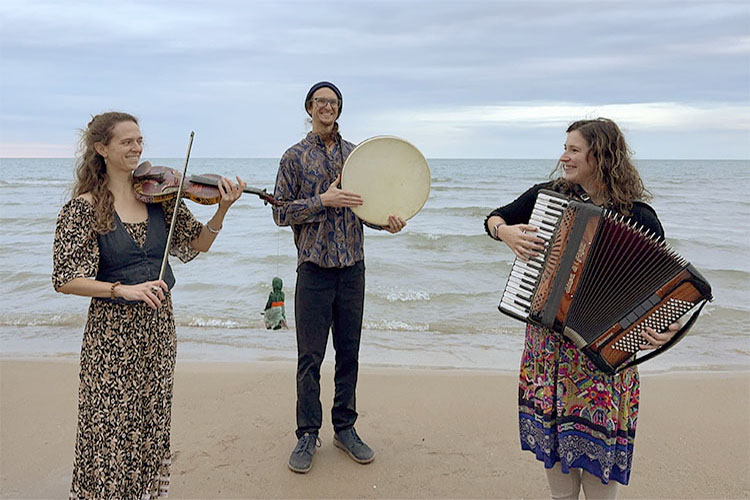 Three musicians playing a fiddle, hand drum, and accordion on a beach.