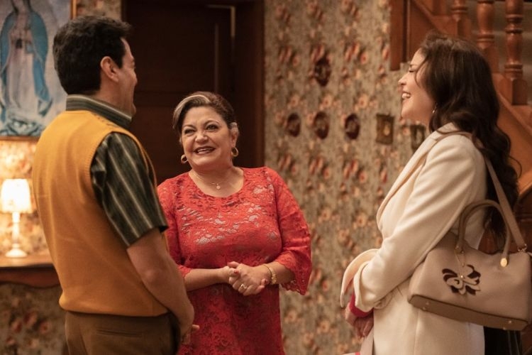 A man and two women stand indoors, smiling and talking in a living room.