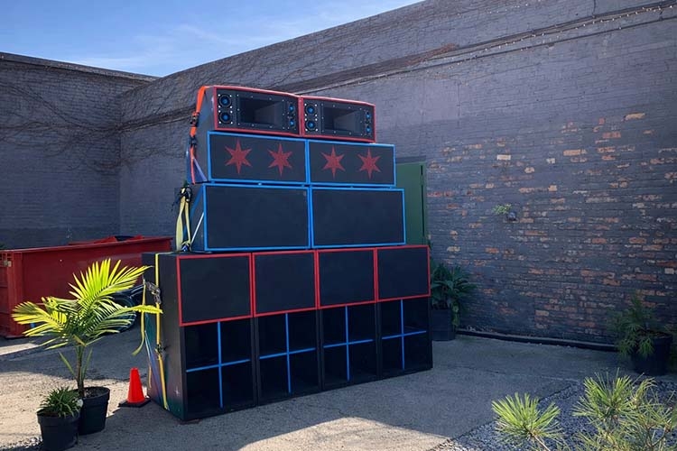 Photograph of a large stack of black sound system speakers outdoors against a brick wall.