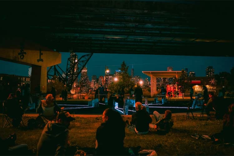 Photograph of people sitting on grass at night viewing a music performance with a light installation.