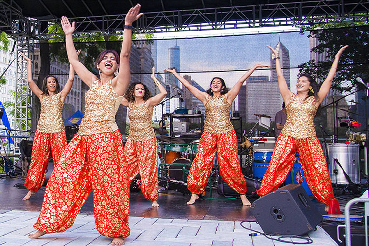 Five women in sparkling gold tops and patterned red pants dance joyfully on an outdoor stage.
