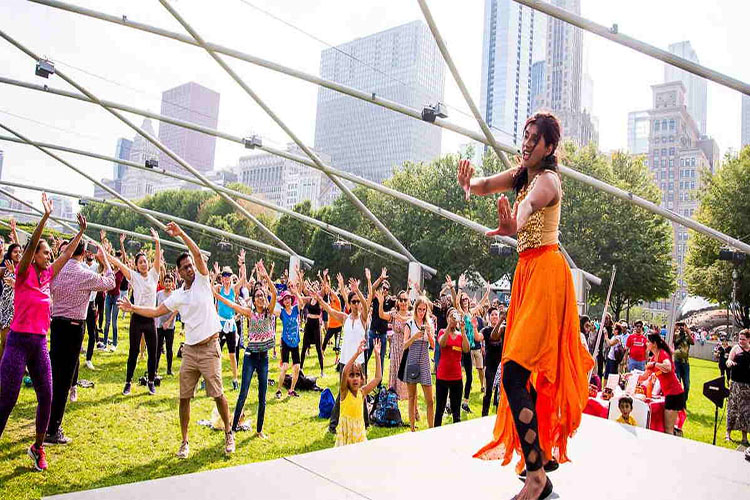A woman in an orange skirt leads a fitness class on a stage in a city park.