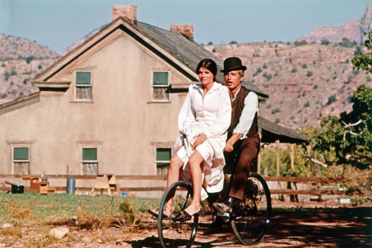 Katharine Ross and Paul Newman ride a bicycle together in front of a rustic house in a photograph.