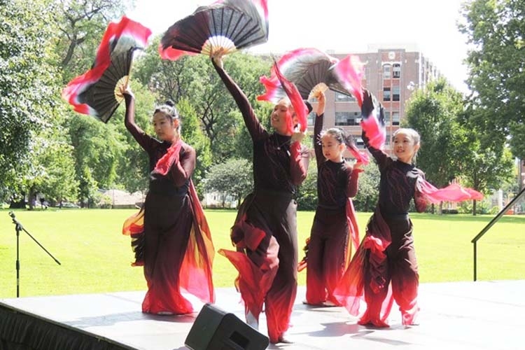 Four dancers in red and black performance outfits holding fans on an outdoor stage.
