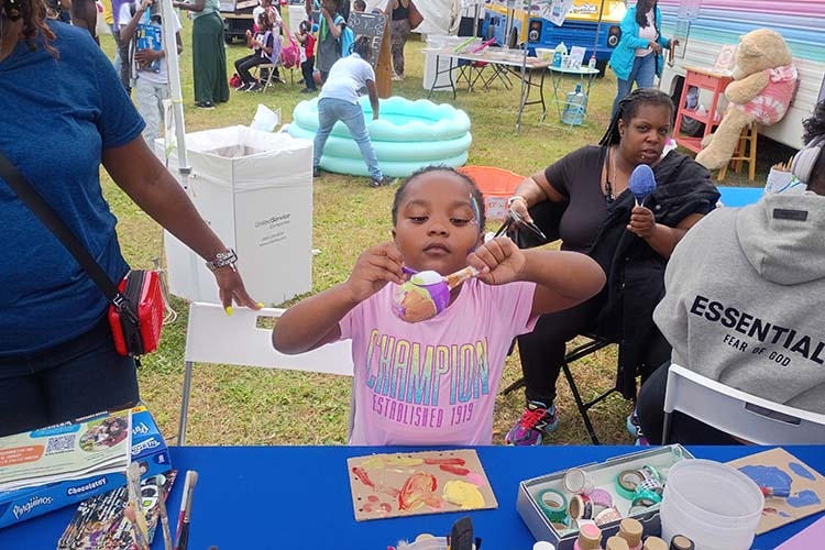 A young child in a pink shirt focuses on painting a small, spherical object at an outdoor event.