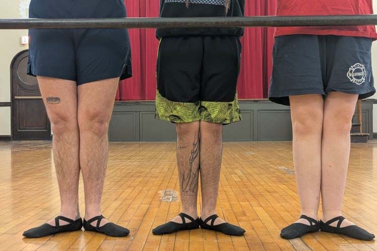 Three people standing in first position at a ballet barre in a studio.