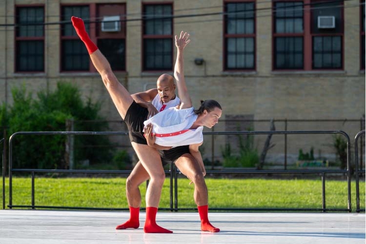 Two dancers in black shorts and red socks perform a choreographed lift outdoors.