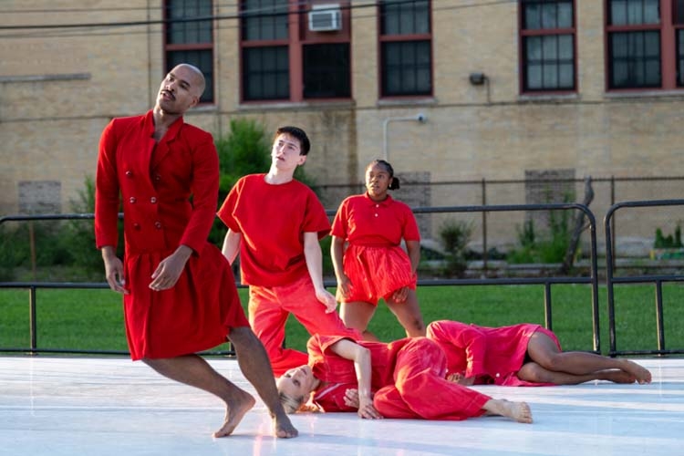 Photograph of dancers in red outfits performing on a white outdoor stage.