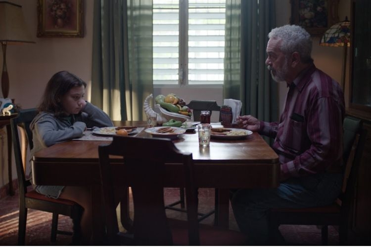 A young girl and an older man sit at a dining table for a quiet meal in a dimly lit room.