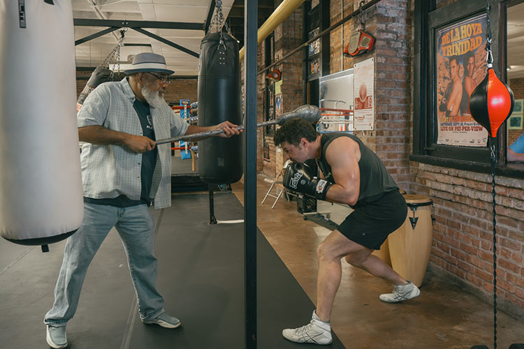 An older trainer in a hat guides a boxer using a weighted mace in a brick-walled boxing gym.