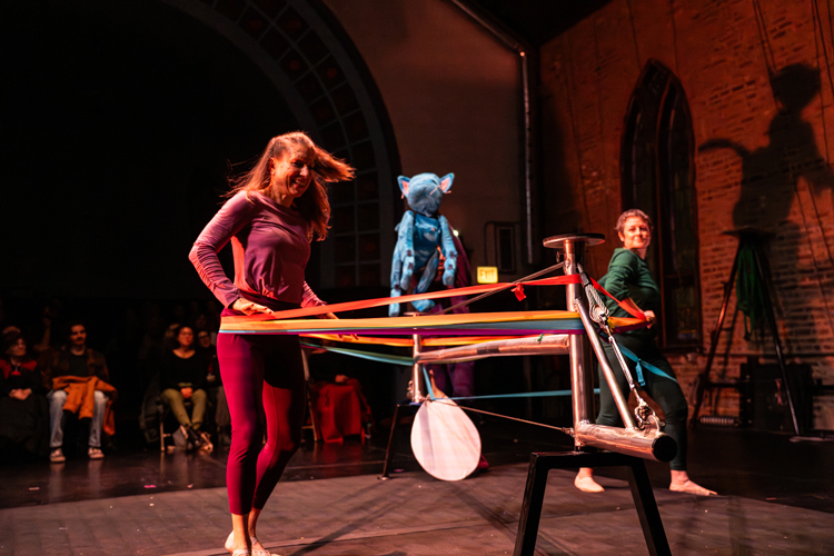 Photograph of two performers and a blue puppet interacting with colorful ribbons on a stage.