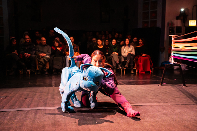A performer in purple kneels on a stage, manipulating a large blue cat puppet for an audience.