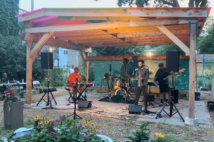 Photograph of a band performing on an outdoor stage under a wooden structure.