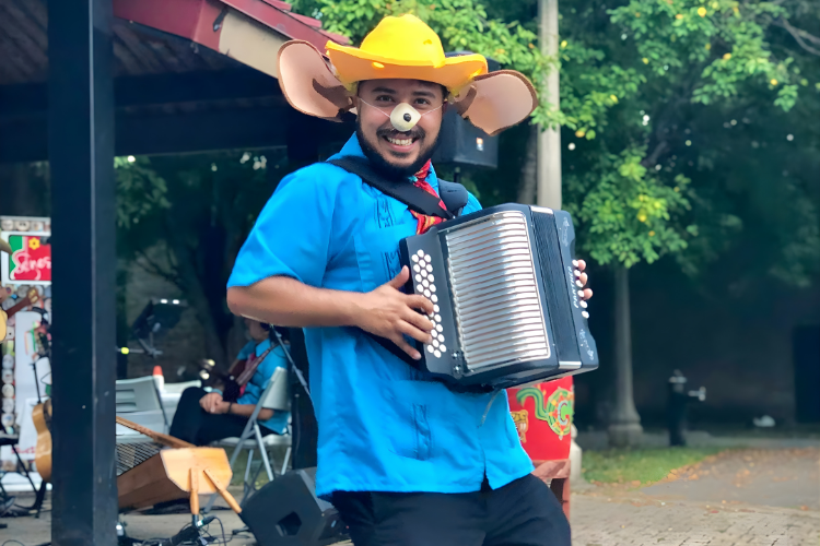 A smiling man wearing a mouse hat and ears plays a button accordion outdoors.