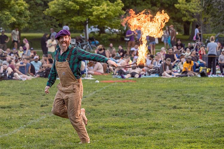 A barefoot fire performer wearing a purple cap and overalls swings a flaming torch in a park.