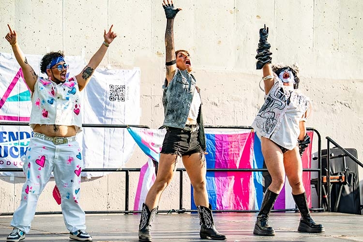 Three performers in theatrical costumes and makeup stand on stage with arms raised.