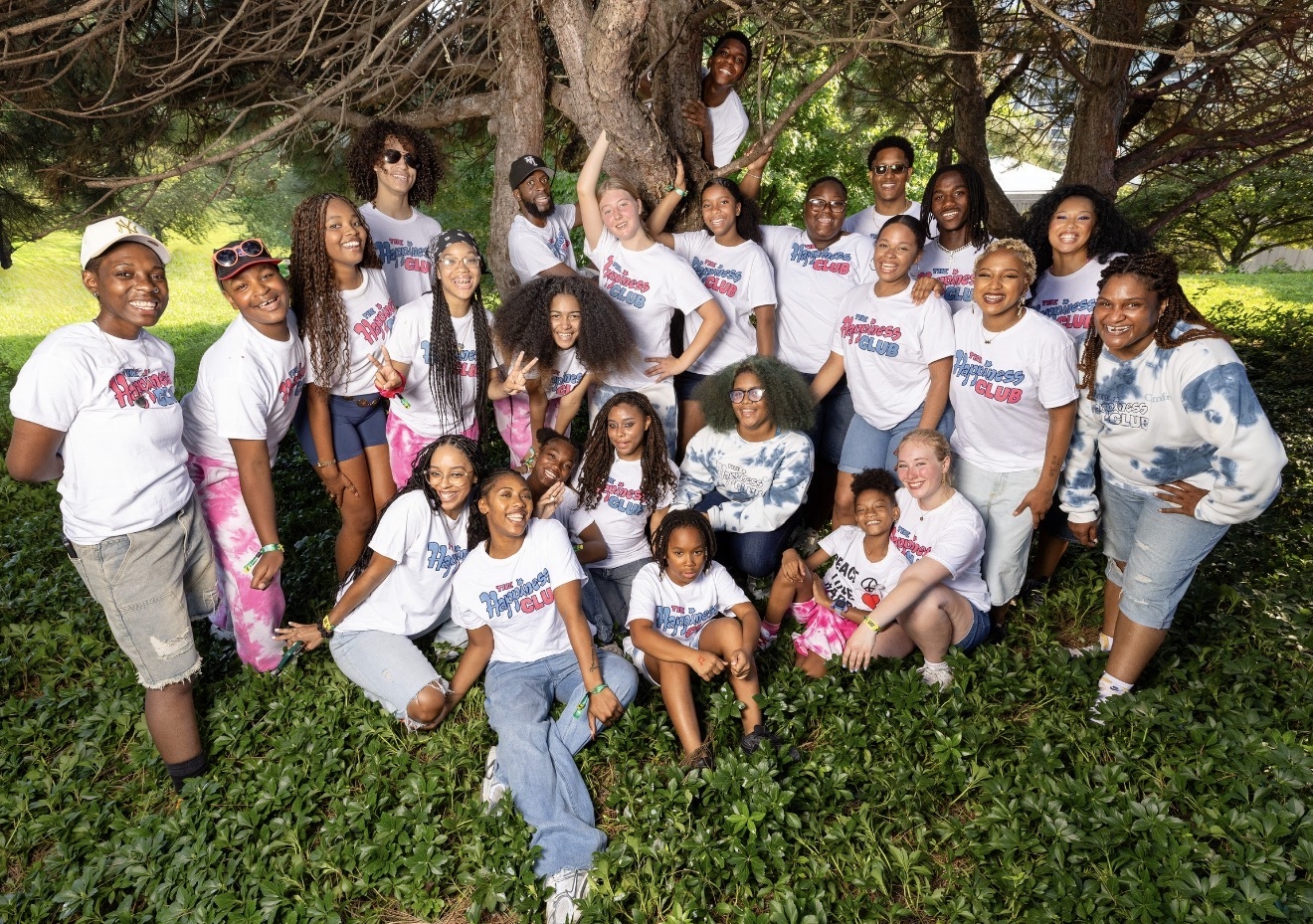 A diverse group of young people wearing "The Happiness Club" shirts posing together outdoors.