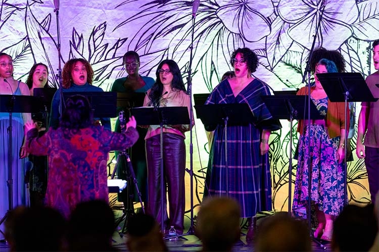 A choir performs on stage in front of a floral backdrop, with a conductor facing them.