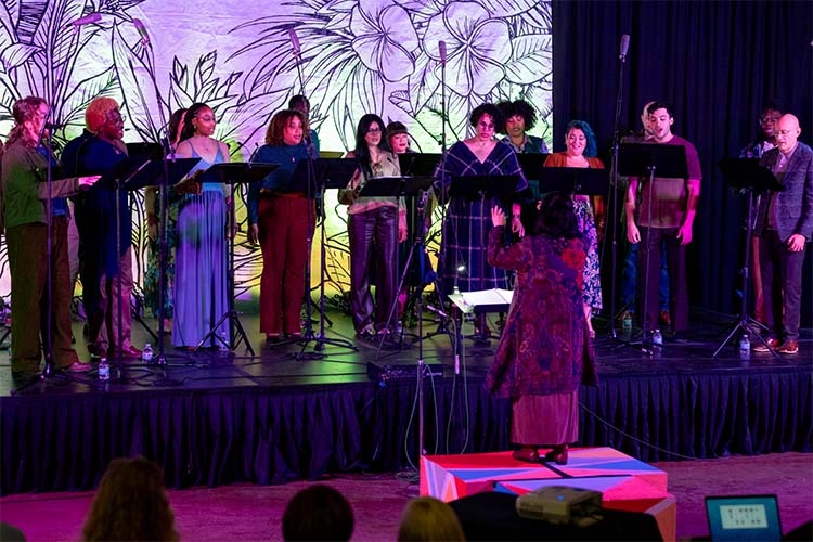 A choir performs on a stage in front of a floral mural, led by a conductor with their back turned.