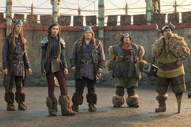 Five Vikings in horned helmets stand in a row against a stone wall.