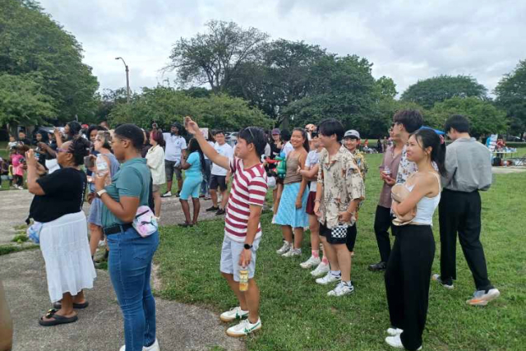 A diverse group of young adults standing in a park, many looking toward an event out of frame.