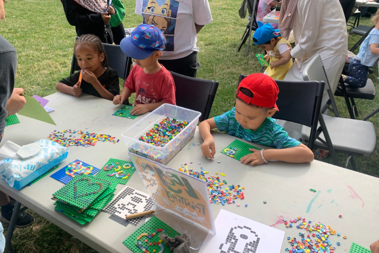 Children sitting at a table outdoors, creating patterns on small green plastic boards with beads.