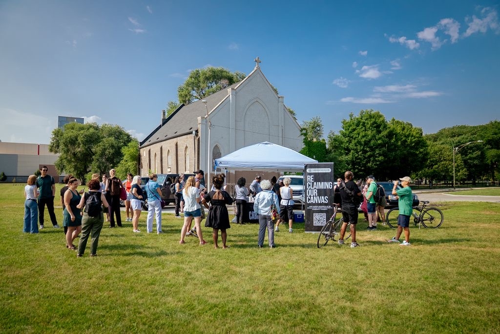 A group of people gathers on a sunny, grassy lawn in front of an old church and an event tent.