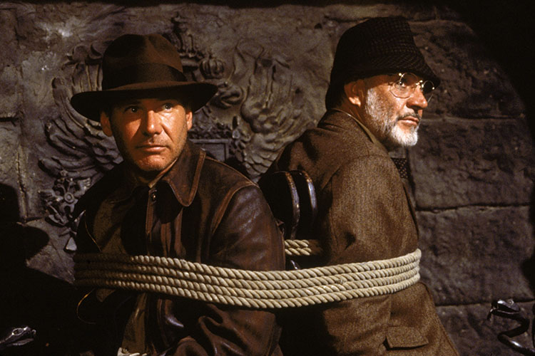Photograph of Indiana Jones and his father tied back-to-back in a dimly lit, stony room.